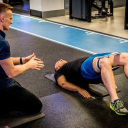 Norbert Gotz - trener personalny Kraków - Trener personalny demonstruje ćwiczenie z gumą na nogach klienta leżącego na macie w siłowni. Ujęcie z boku.