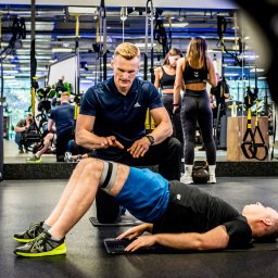 Norbert Gotz - trener personalny Kraków - Trener nadzoruje ćwiczenie mostu biodrowego z gumą oporową w siłowni. W tle osoby i sprzęt do ćwiczeń. Dynamiczne ujęcie z bliska.