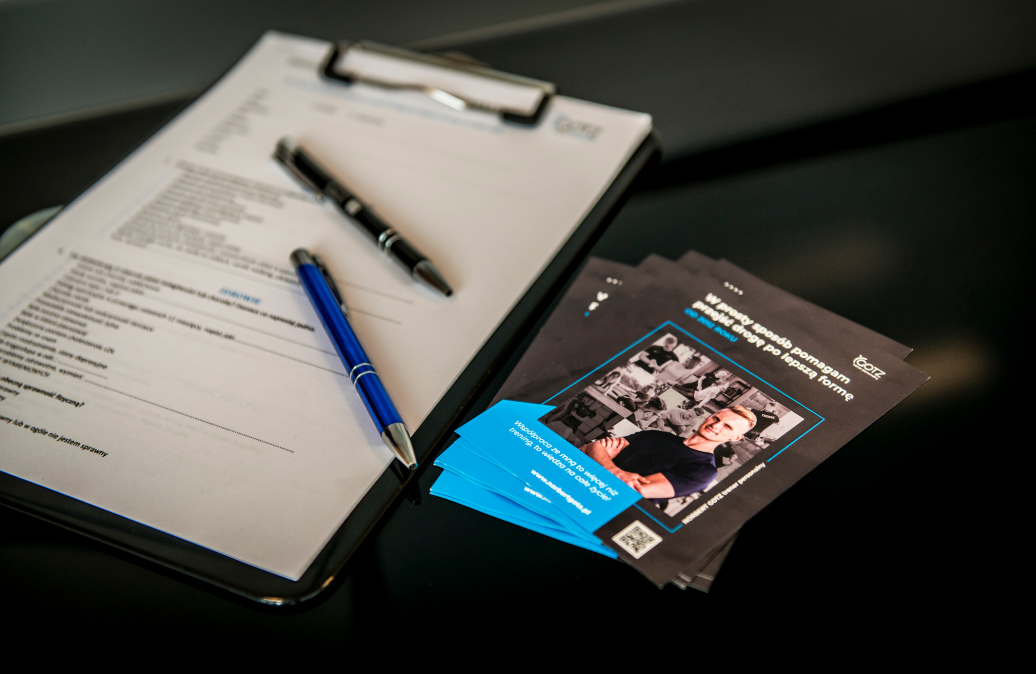 Formularz, wizytówki i ulotki trenera personalnego na błyszczącym, czarnym stole. Widoczne długopisy. Ulotka z wizerunkiem trenera i hasłem o lepszej formie.