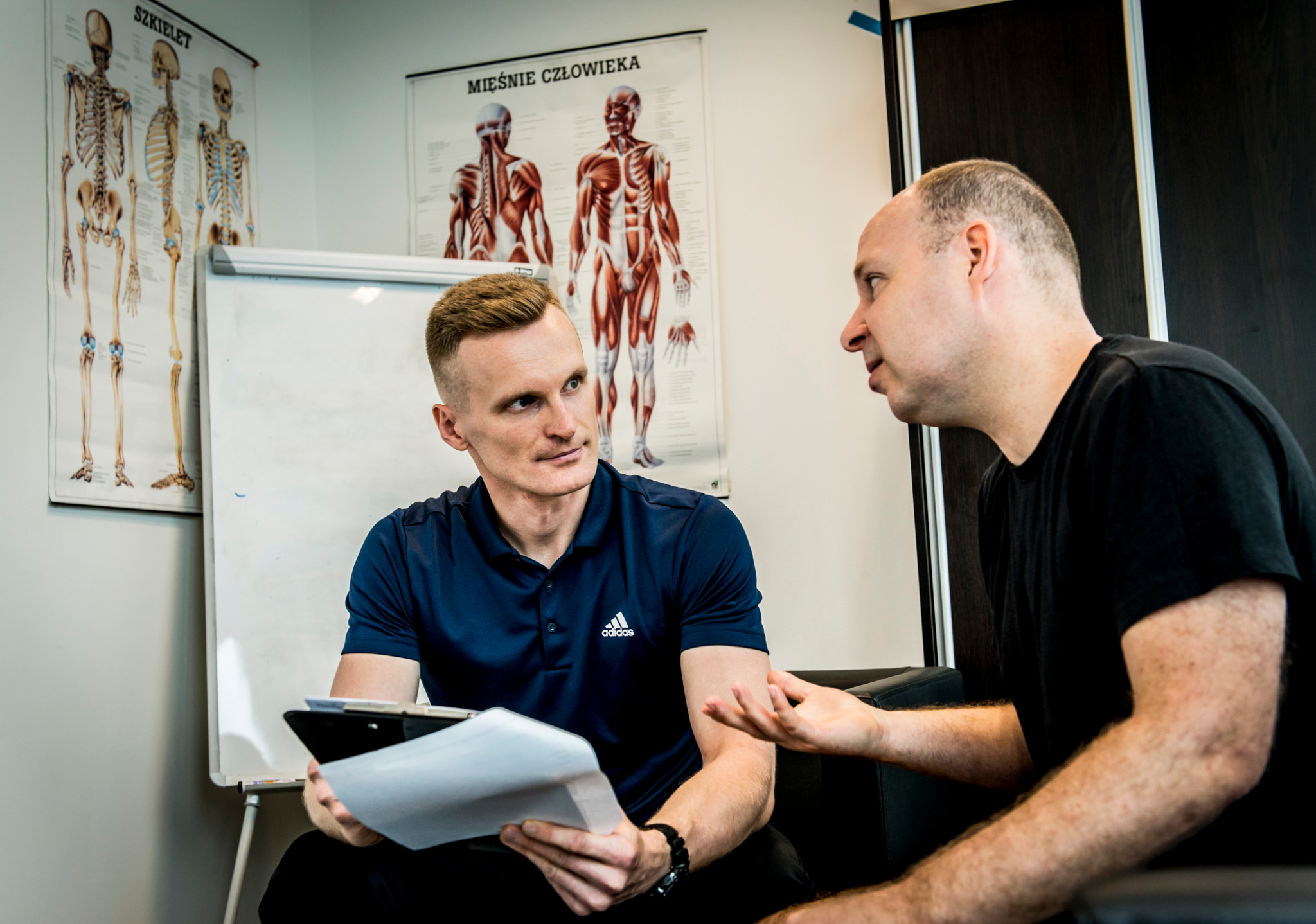 Trener personalny omawia plan treningowy z klientem, w tle plansze anatomiczne. Mężczyźni siedzą na czarnej sofie, trener trzyma dokumenty na podkładce.