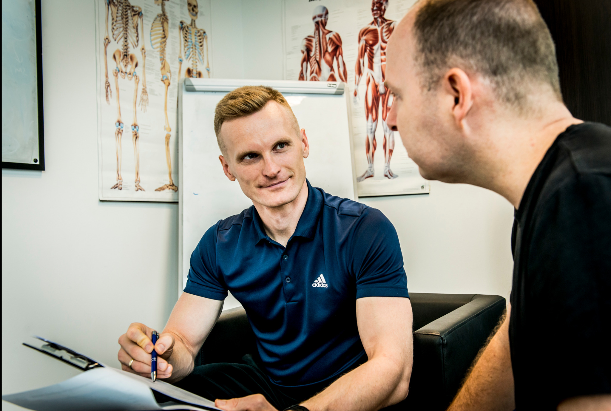 Trener personalny w granatowej koszulce Adidas omawia plan treningowy z klientem, w tle plansze anatomiczne i tablica suchościeralna.