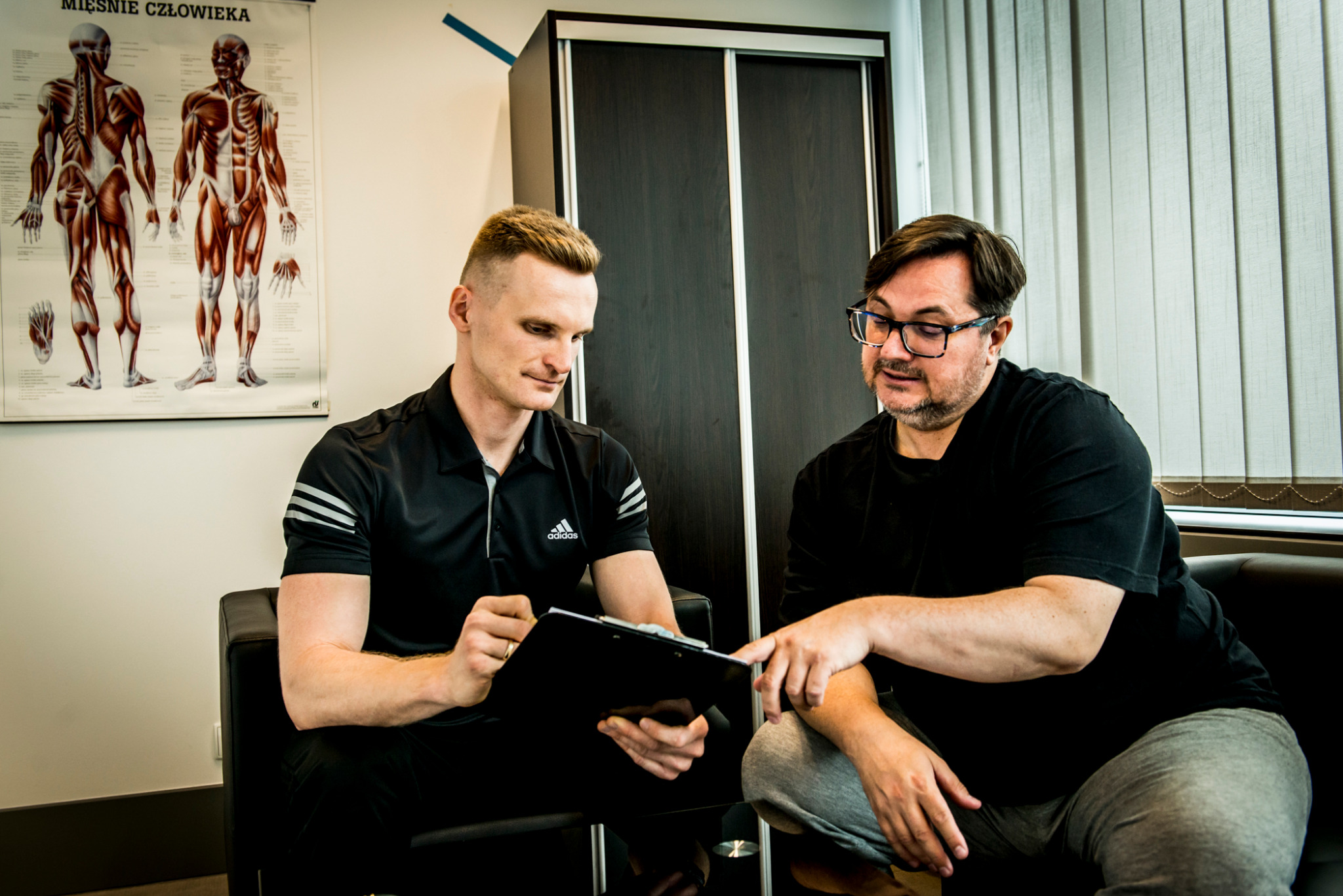 Konsultacja trenera personalnego z klientem w gabinecie. Mężczyźni siedzą na kanapie, trener omawia plan treningowy, wskazując na dokument na podkładce.