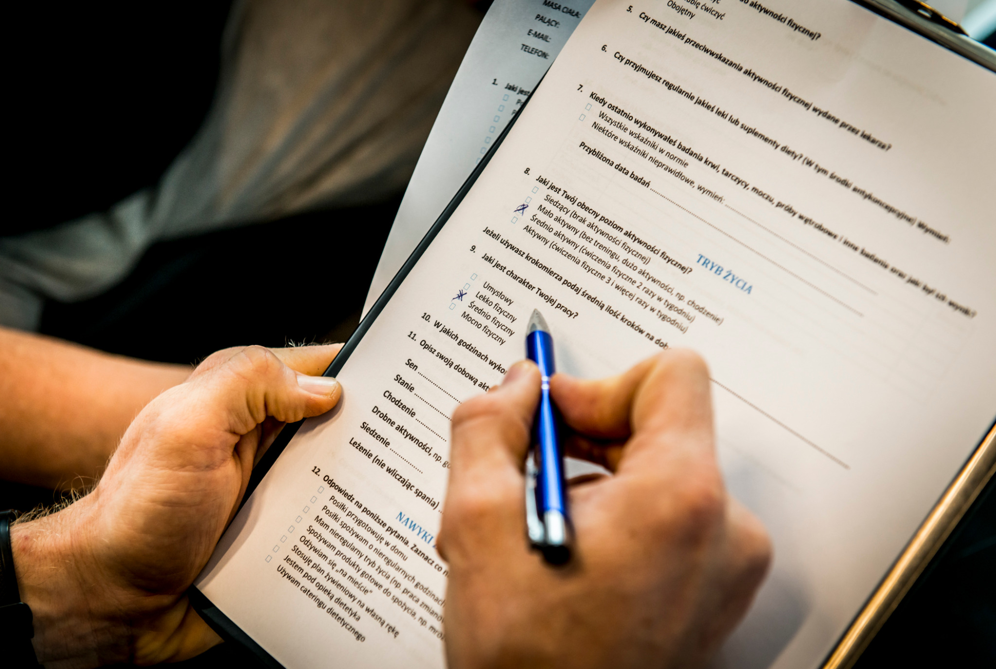 Wypełnianie kwestionariusza dotyczącego aktywności fizycznej i trybu życia, niebieski długopis w dłoni zaznacza odpowiedź. Formularz na białym tle.
