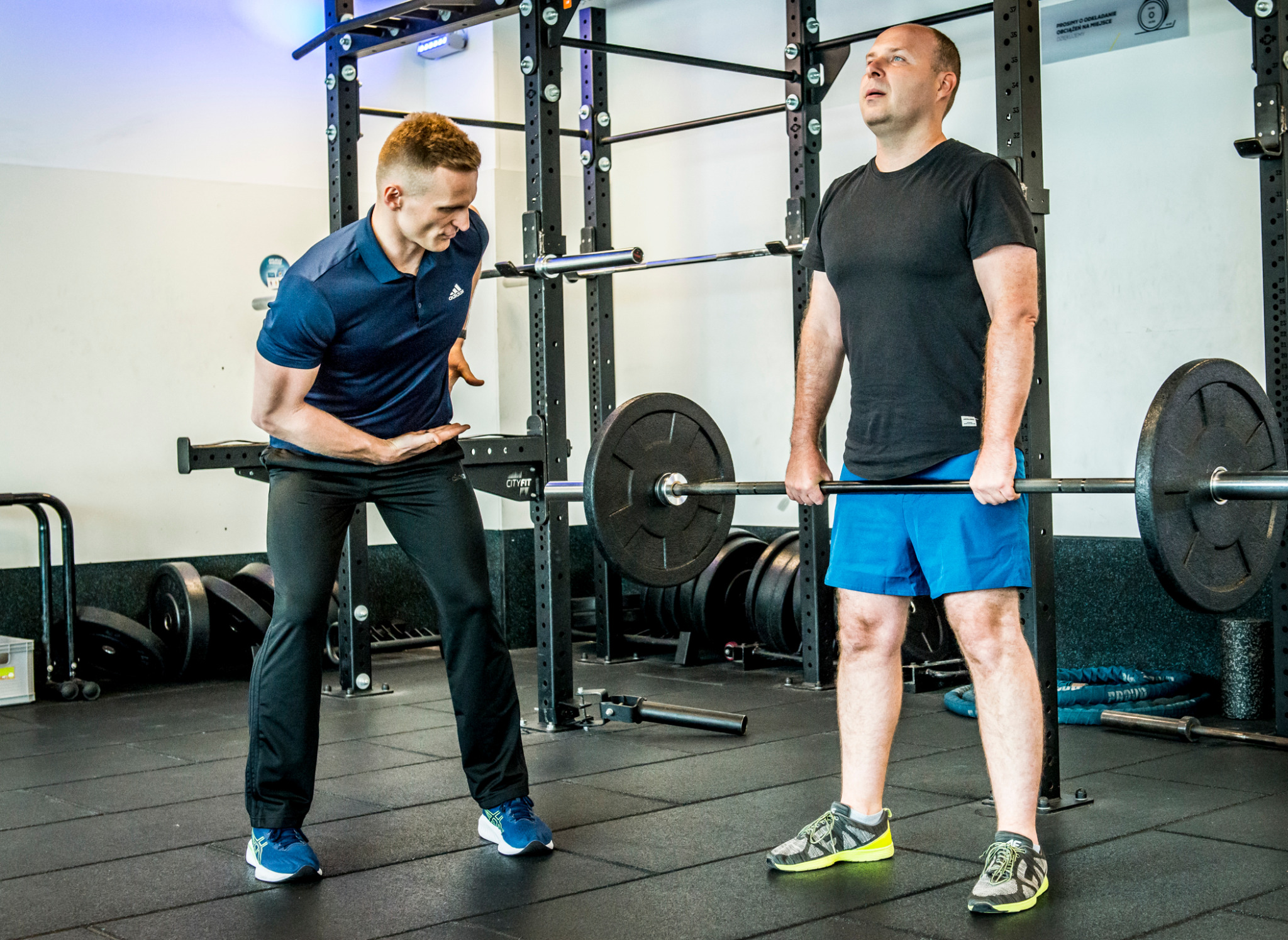 Trener demonstruje prawidłową postawę podczas podnoszenia ciężarów klientowi w siłowni z czarną podłogą i sprzętem do ćwiczeń siłowych.