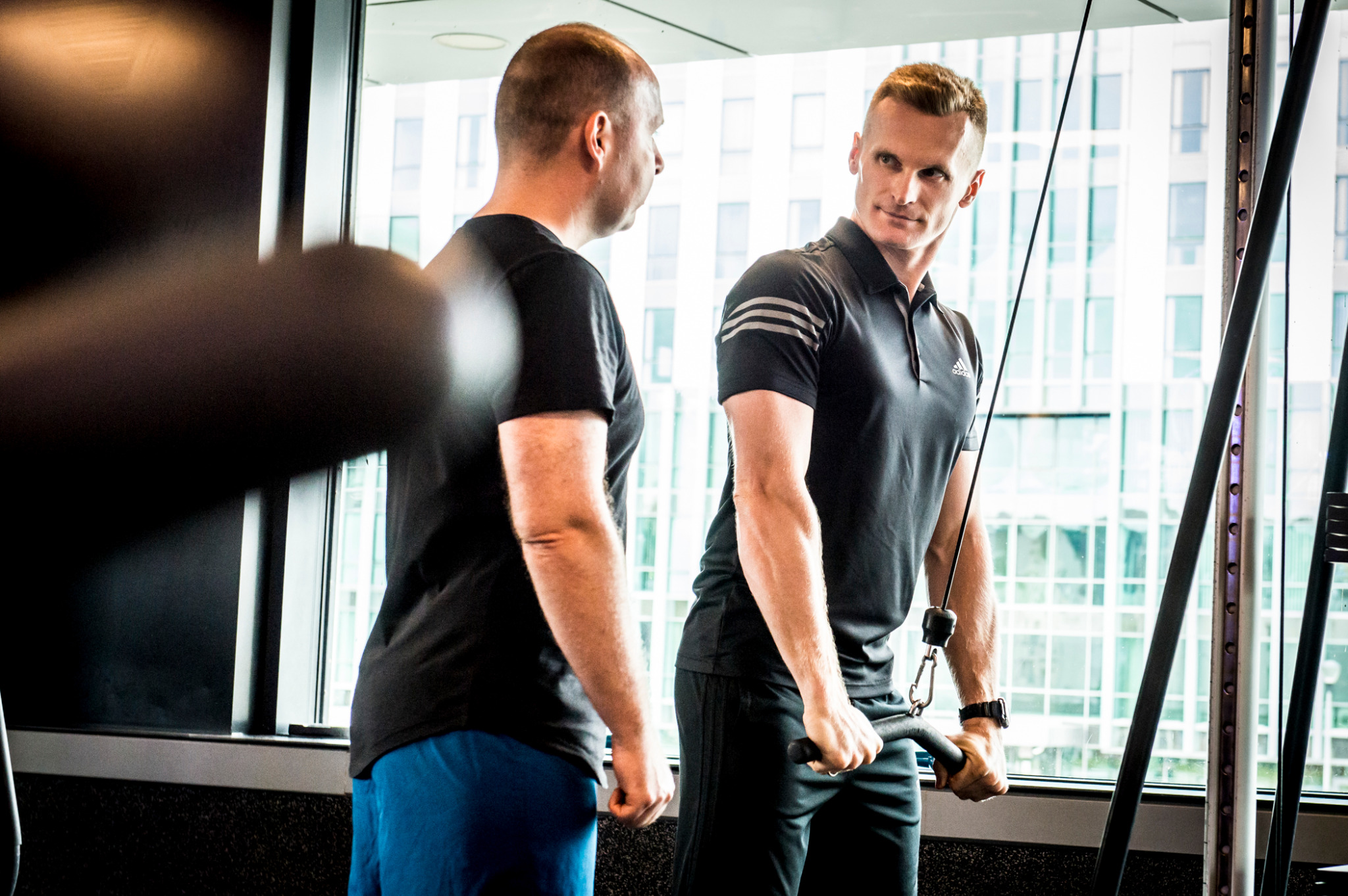 Mężczyzna w czarnej koszulce ćwiczy na maszynie w siłowni, pod okiem trenera. Za nimi okno z widokiem na budynki.