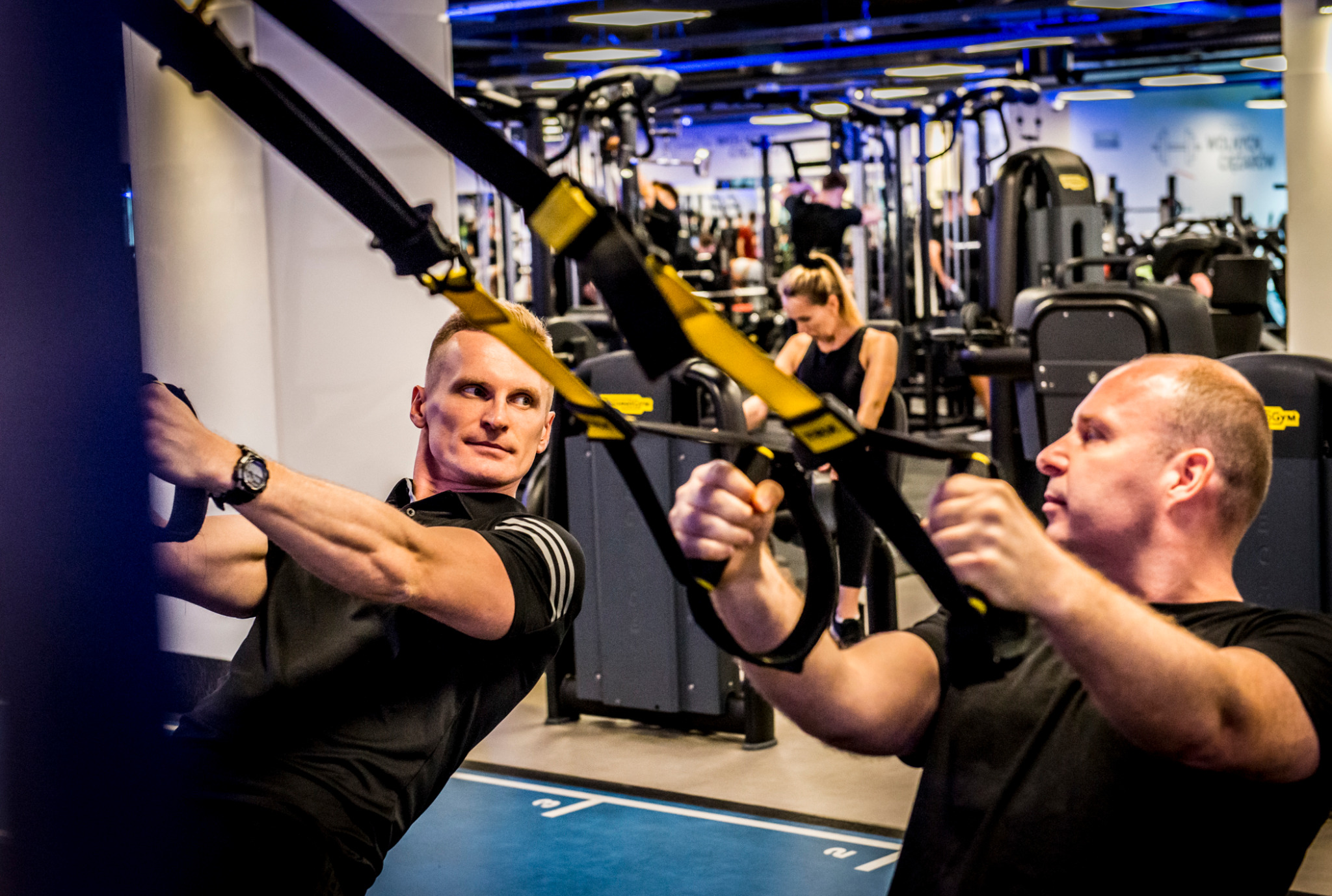 Mężczyźni ćwiczą na TRX w siłowni, w tle widoczny sprzęt do ćwiczeń i inne osoby trenujące. Jeden z mężczyzn ma zegarek na lewej ręce.