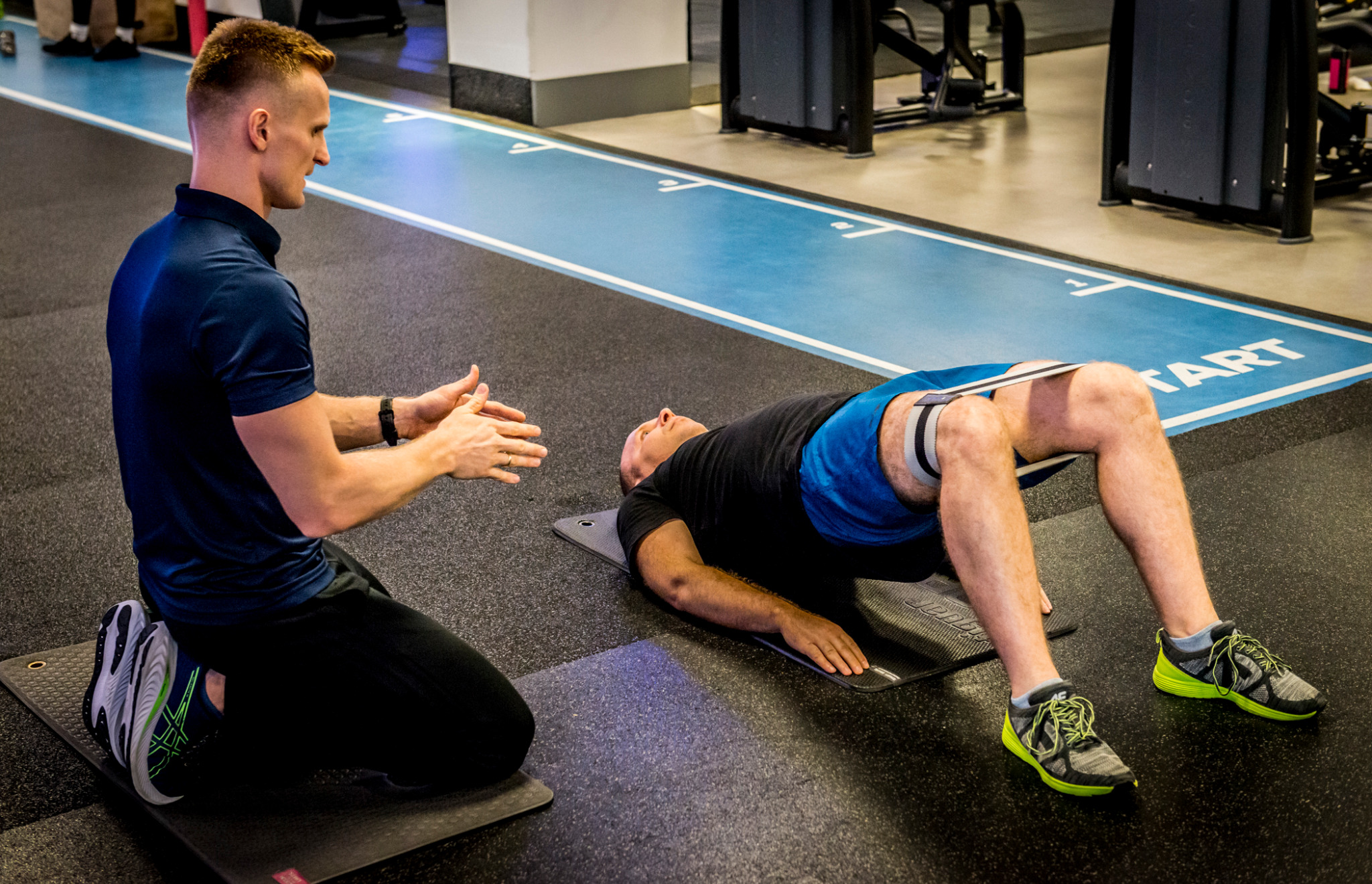 Trener personalny demonstruje ćwiczenie z gumą na nogach klienta leżącego na macie w siłowni. Ujęcie z boku.