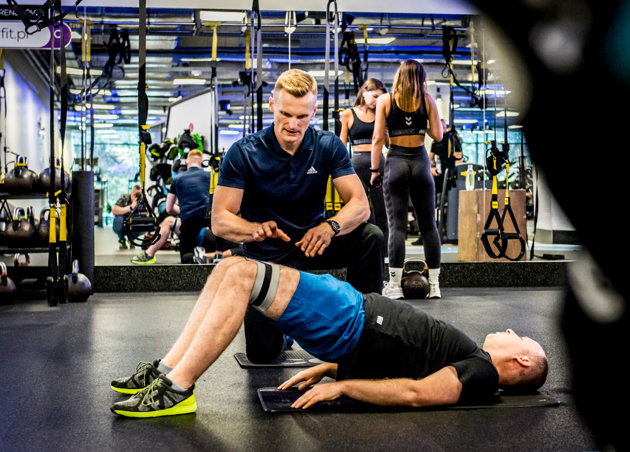 Trener nadzoruje ćwiczenie mostu biodrowego z gumą oporową w siłowni. W tle osoby i sprzęt do ćwiczeń. Dynamiczne ujęcie z bliska.