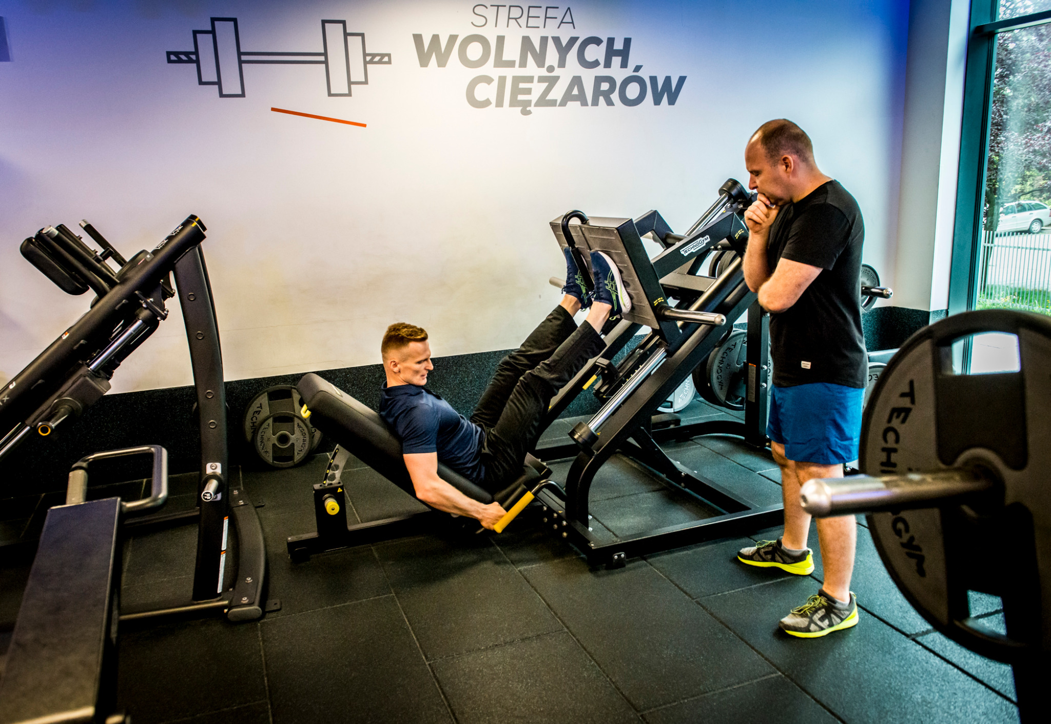 Mężczyzna ćwiczy na maszynie do wyciskania nóg w siłowni, pod okiem trenera. W tle widoczny napis 'Strefa wolnych ciężarów'.