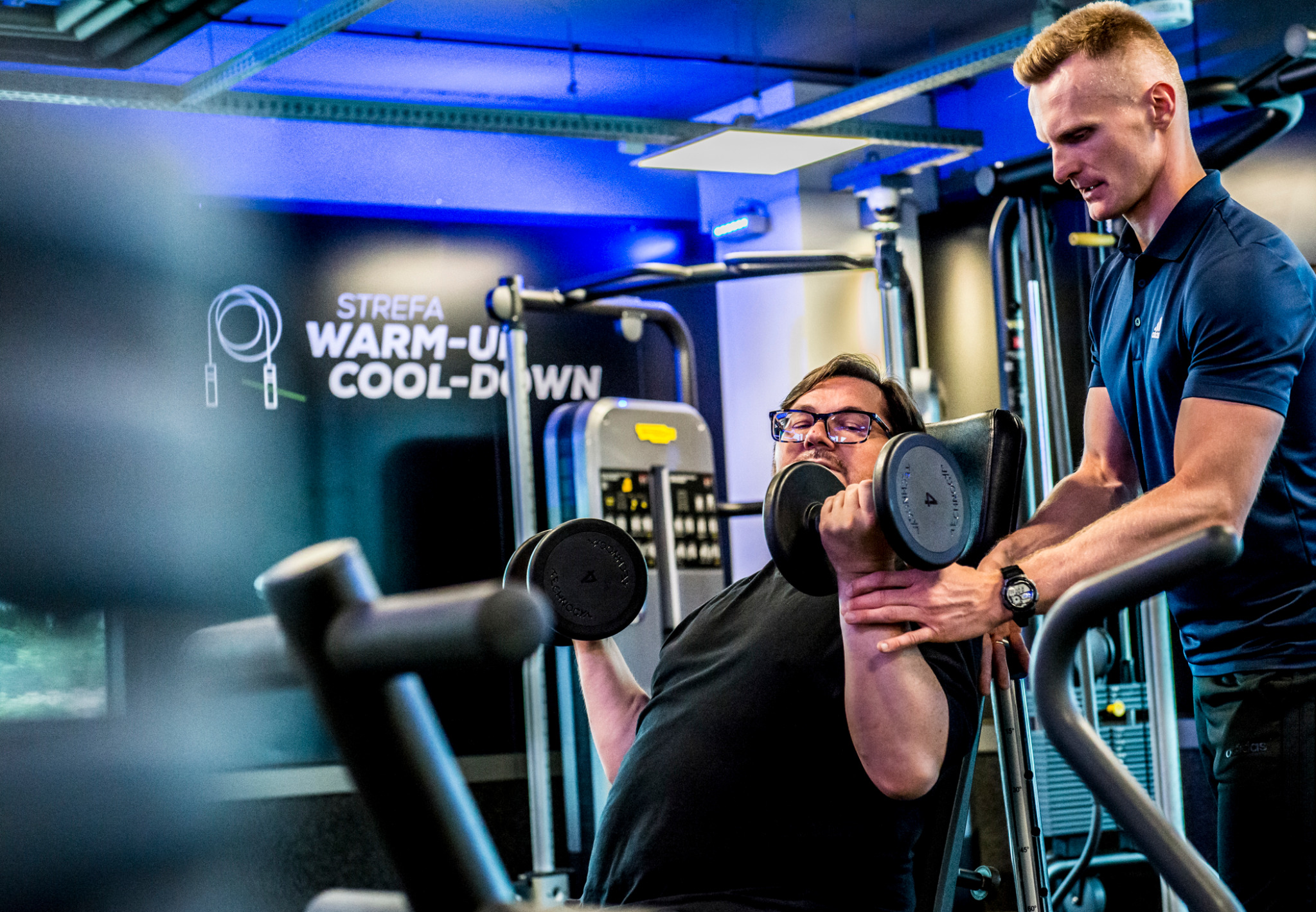 Mężczyzna ćwiczy z hantlami pod okiem trenera w nowoczesnej siłowni z napisem 'Strefa Warm-Up/Cool-Down' w tle, co sugeruje przygotowanie lub zakończenie treningu.