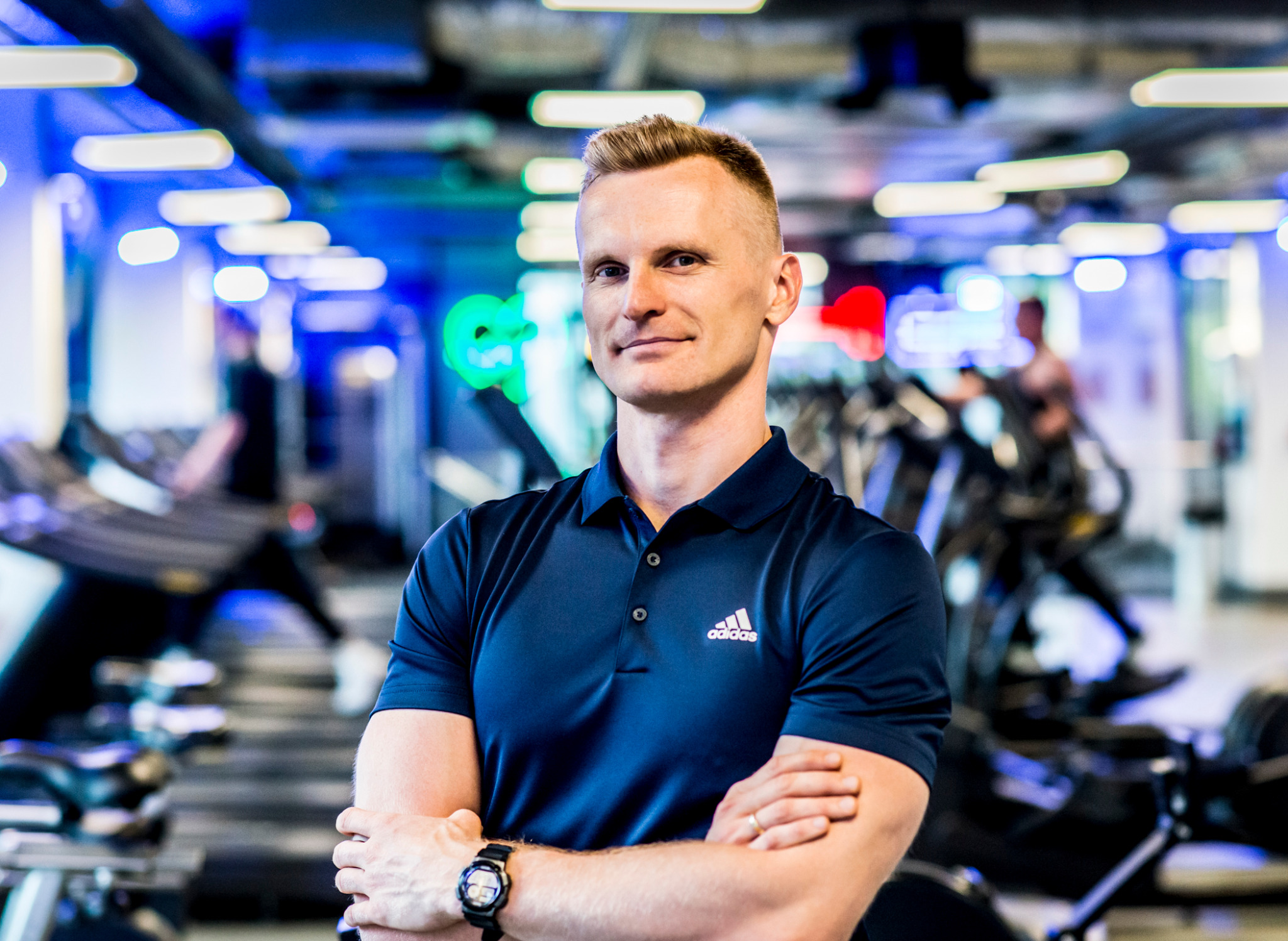 Mężczyzna w sportowej koszulce polo na tle siłowni z oświetleniem LED. Skrzyżowane ręce, zegarek na nadgarstku. Sprzęt do ćwiczeń w tle.