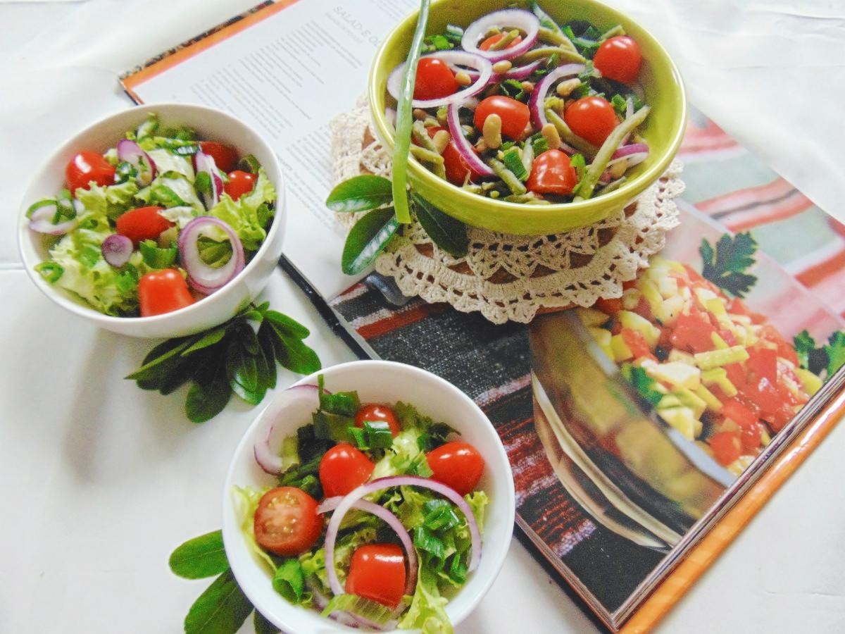 Trzy miseczki sałatek z pomidorami, czerwoną cebulą i zieloną sałatą, ustawione na białym tle obok otwartej książki kucharskiej z przepisem na sałatkę.