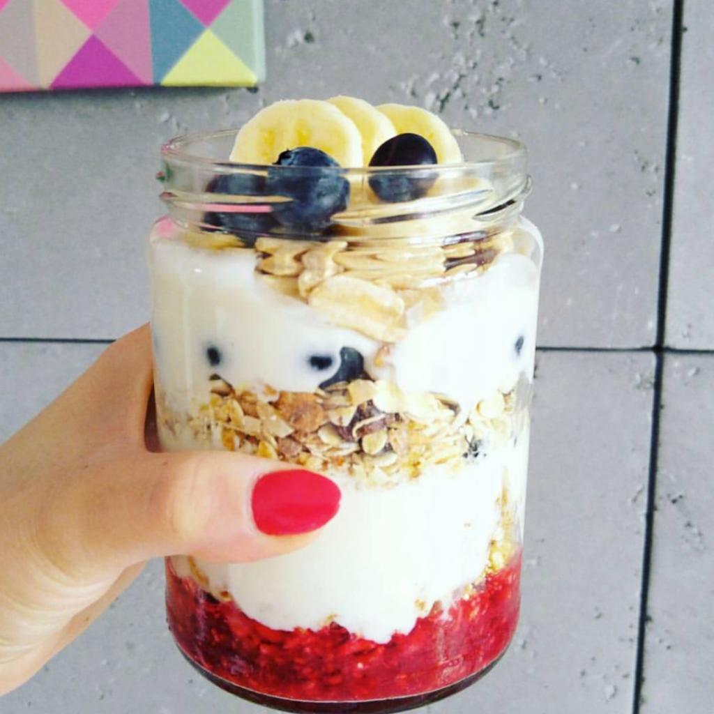 Szklany słoik z warstwami jogurtu, musli, owoców leśnych, banana i borówek, trzymany w dłoni z czerwonym lakierem na paznokciach na tle szarej ściany.