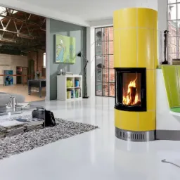 Żółty, ceramiczny piec kominkowy z paleniskiem, płomienie widoczne przez szybę, na tle industrialnego wnętrza z dużymi oknami i widokiem na budynki portowe. Obok pieca zielony, designerski fotel.