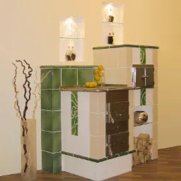 Tradycyjny piec kaflowy z zielonymi akcentami, uzupełniony dekoracyjną wazą z suchymi gałązkami i owocami na blacie, w tle wnęki ścienne z ozdobnymi butelkami.