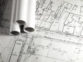 Zrolowane plany architektoniczne leżące na rozłożonym szczegółowym rysunku technicznym z zaznaczonymi budynkami i numerami działek.