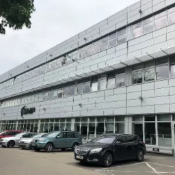 Nowoczesny budynek biurowo-usługowy z szarą elewacją panelową i rzędem zaparkowanych samochodów na pierwszym planie, widoczne logo firmy na fasadzie.