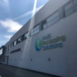 Zewnętrzny widok hali produkcyjno-magazynowej z logo Multi Packaging Solutions na ścianie, widoczne okna i fragment infrastruktury komunikacyjnej, błękitne niebo z smugami kondensacyjnymi.
