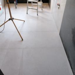 Mir-pol - Podłoga i ściany w trakcie wykańczania w łazience, widoczne płytki w odcieniach szarości, drabina malarska, lampa budowlana na trójnogu i wystające rury instalacji hydraulicznej.