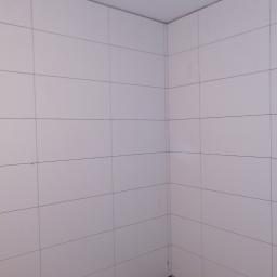 Mir-pol - Narożnik wykończonej łazienki z białymi, kwadratowymi płytkami ceramicznymi ułożonymi w regularny wzór, widoczne fugi.