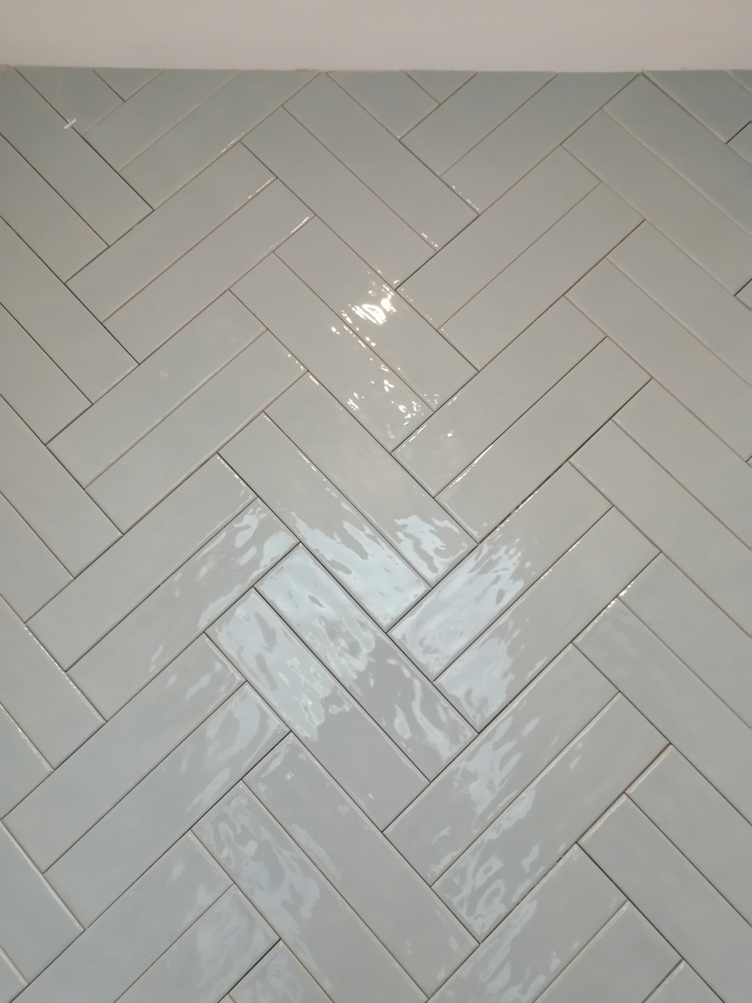 Ściana wyłożona błyszczącymi, podłużnymi, szarymi płytkami ułożonymi w jodełkę, widoczne odbicia światła.