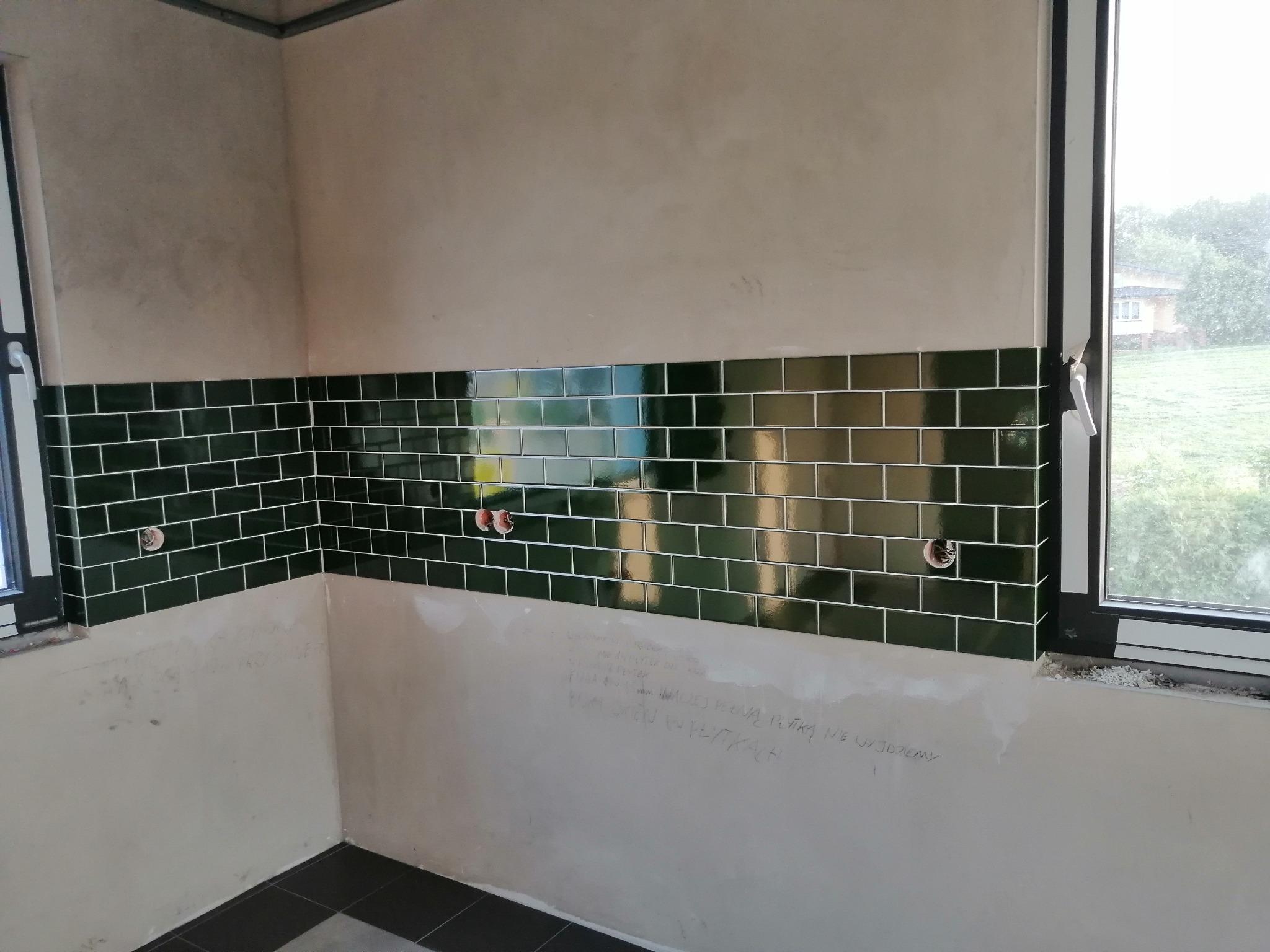 Fragment ściany z położonymi ciemnozielonymi, błyszczącymi płytkami w układzie cegiełki, widoczne otwory pod instalacje, ściana częściowo przygotowana do dalszych prac wykończeniowych, okno...