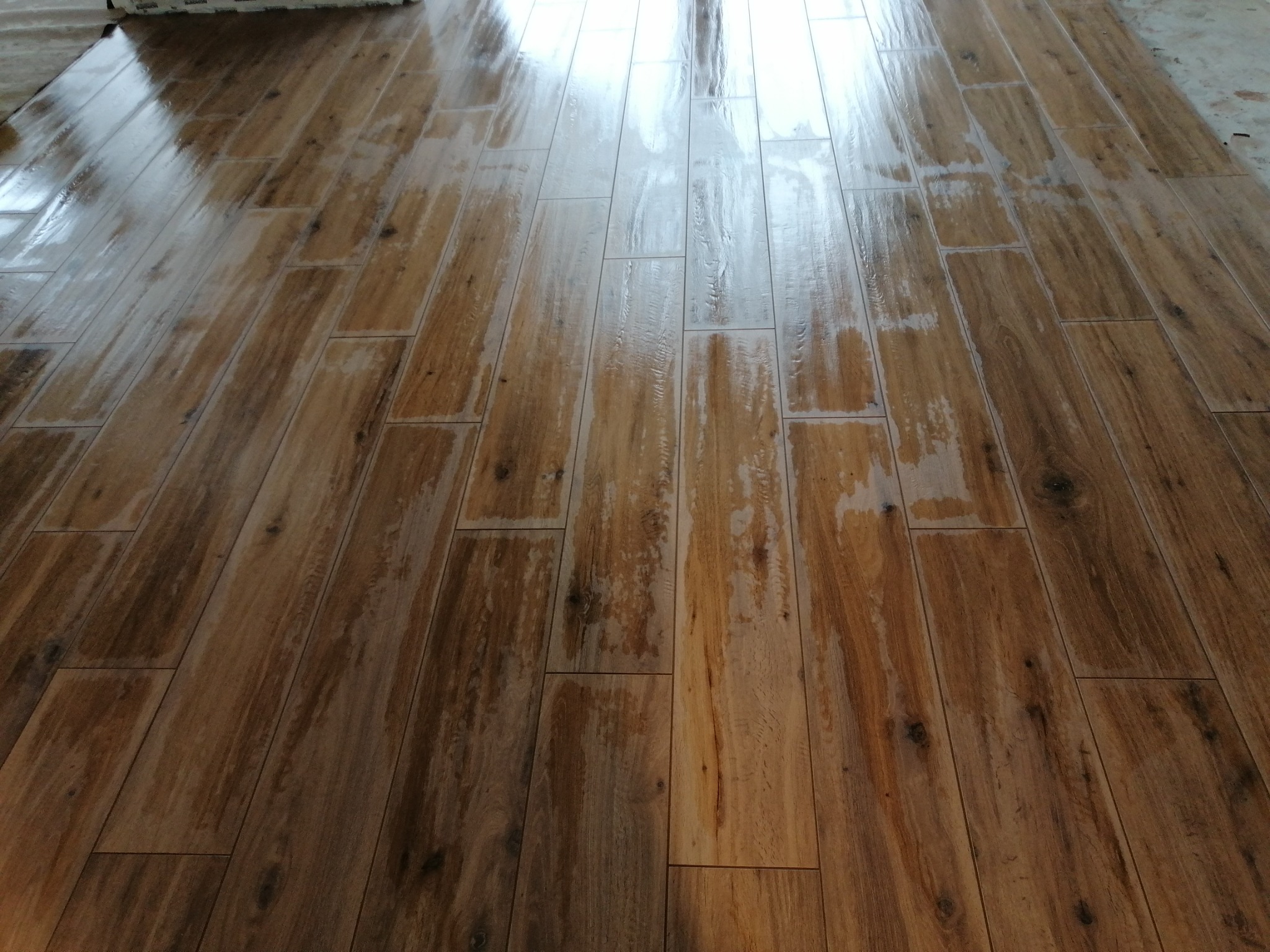 Świeżo ułożona, błyszcząca podłoga z płytek imitujących drewno, z widocznymi refleksami światła i fugami, w trakcie czyszczenia lub schnięcia.