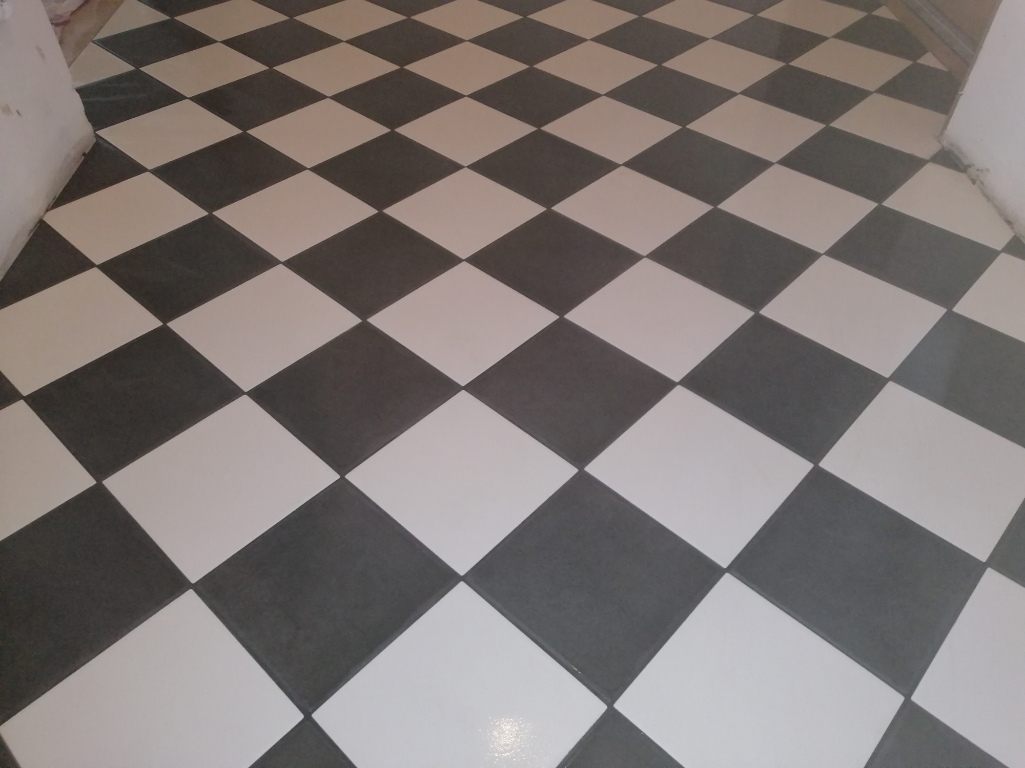 Ułożona podłoga z płytek w szachownicę, biało-szara, widok z góry, fuga jasnoszara, ściana w tle nie wykończona.