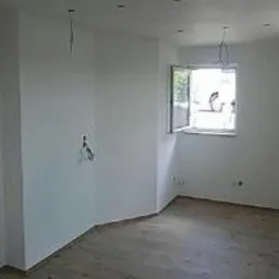 Jasne wnętrze pokoju po remoncie, z drewnianą podłogą ułożoną w jodełkę, świeżo pomalowanymi na biało ścianami, oknem z widokiem na zieleń i wystającymi przewodami elektrycznymi.