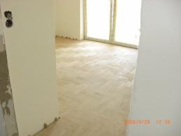 Jasny parkiet ułożony w jodełkę francuską w nowym pomieszczeniu, widoczne niedokończone wykończenia ścian i okna w tle.