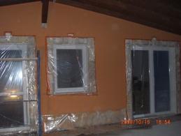 Pomarańczowa ściana budynku z trzema oknami zabezpieczonymi folią malarską, widoczne drewniane elementy konstrukcyjne dachu, ujęcie z dołu w słabym oświetleniu.