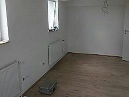 Puste, jasne pomieszczenie po remoncie z drewnianą podłogą, białymi ścianami i grzejnikami. Na podłodze leży metalowa walizka narzędziowa. Widoczne ślady po instalacjach elektrycznych na ścianie.