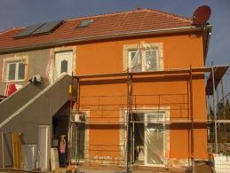 Dwukondygnacyjny budynek w trakcie renowacji elewacji: część ściany otynkowana na pomarańczowo, druga część w szarym tynku, widoczne rusztowania, okna zabezpieczone folią, panele solarne na dachu.