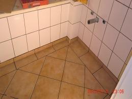 Niedokończona łazienka z białymi, kwadratowymi płytkami ściennymi i brązowymi, większymi płytkami podłogowymi ułożonymi w romby. Widoczny fragment instalacji wodnej i fragment drzwi.