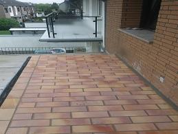 Ułożona z cegieł klinkierowych powierzchnia tarasu na dachu, widok z góry, z balustradą w tle i fragmentem elewacji budynku z cegły.