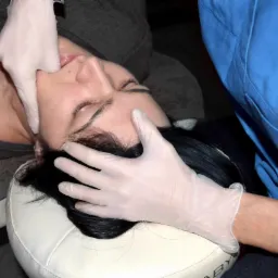 Kobieta leżąca na kozetce podczas terapii manualnej twarzy i szyi, wykonywanej przez terapeutę w rękawiczkach ochronnych.