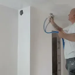 Pracownik w Tyszowcach aplikuje gładź natryskową na sufit, używając pistoletu pneumatycznego podłączonego niebieskim przewodem, widoczny fragment ściany z nierównościami przed obróbką.