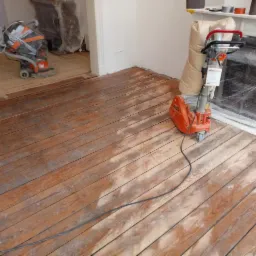 Szlifowanie drewnianej podłogi parkietowej za pomocą szlifierki podłogowej, widoczny fragment podłogi przed i po szlifowaniu, zabezpieczony kominek w tle.