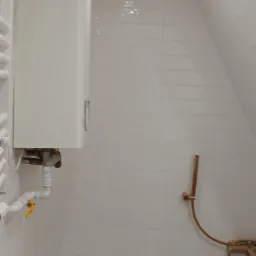 Biała łazienka na poddaszu z grzejnikiem, bojlerem i baterią prysznicową w kolorze miedzianym. Minimalistyczny design, jasne płytki, skosy.