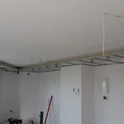 Montaż metalowej konstrukcji sufitu podwieszanego w jasnym pomieszczeniu, widoczne profile i przewody elektryczne.