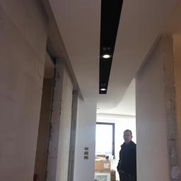 Prace Wykończeniowe Krzysztof Mleczko - Wnętrze w trakcie prac wykończeniowych: sufit z czarnym pasem i oświetleniem punktowym, surowe ściany, widoczny fragment pomieszczenia z oknem i mężczyzna w tle.