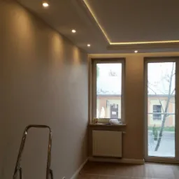 Pokój po malowaniu ścian na kolor beżowy, widoczne listwy sufitowe z oświetleniem LED, zabezpieczona podłoga i drabina.