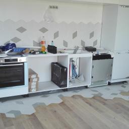 Prace Wykończeniowe Krzysztof Mleczko - Montaż mebli kuchennych w trakcie wykończenia wnętrza: widoczne białe szafki bez frontów, sprzęt AGD w zabudowie, płytki podłogowe heksagonalne częściowo ułożone na drewnianej podłodze,...