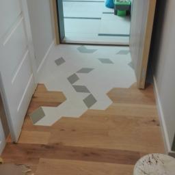 Prace Wykończeniowe Krzysztof Mleczko - Połączenie drewnianej podłogi i heksagonalnych płytek w odcieniach szarości, widok z perspektywy wnętrza, z widocznym fragmentem białego stołka.