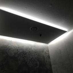 Prace Wykończeniowe Krzysztof Mleczko - Podwieszany sufit z oświetleniem LED w łazience, heksagonalne płytki na ścianie z wzorem patchworkowym, widoczny fragment wentylacji.