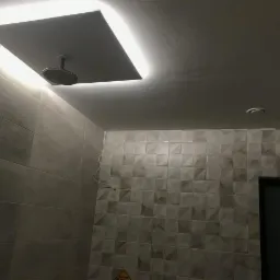 Nowoczesna łazienka z szarymi płytkami o zróżnicowanej fakturze, podwieszanym sufitem z oświetleniem LED i deszczownicą. Widoczne detale wykończenia i instalacji.