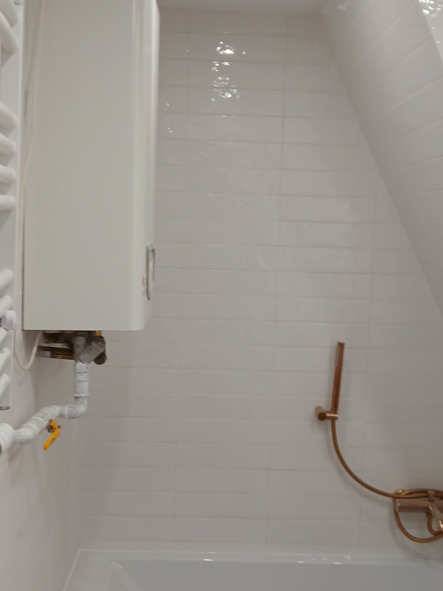 Biała łazienka na poddaszu z grzejnikiem, bojlerem i baterią prysznicową w kolorze miedzianym. Minimalistyczny design, jasne płytki, skosy.