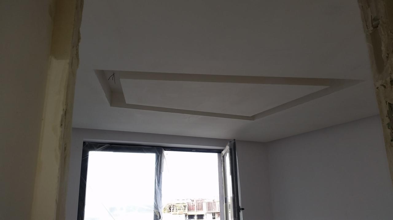 Surowy stan wnętrza z widocznym oknem balkonowym i fragmentem sufitu z obniżeniem w kształcie prostokąta, ściany w trakcie prac wykończeniowych.