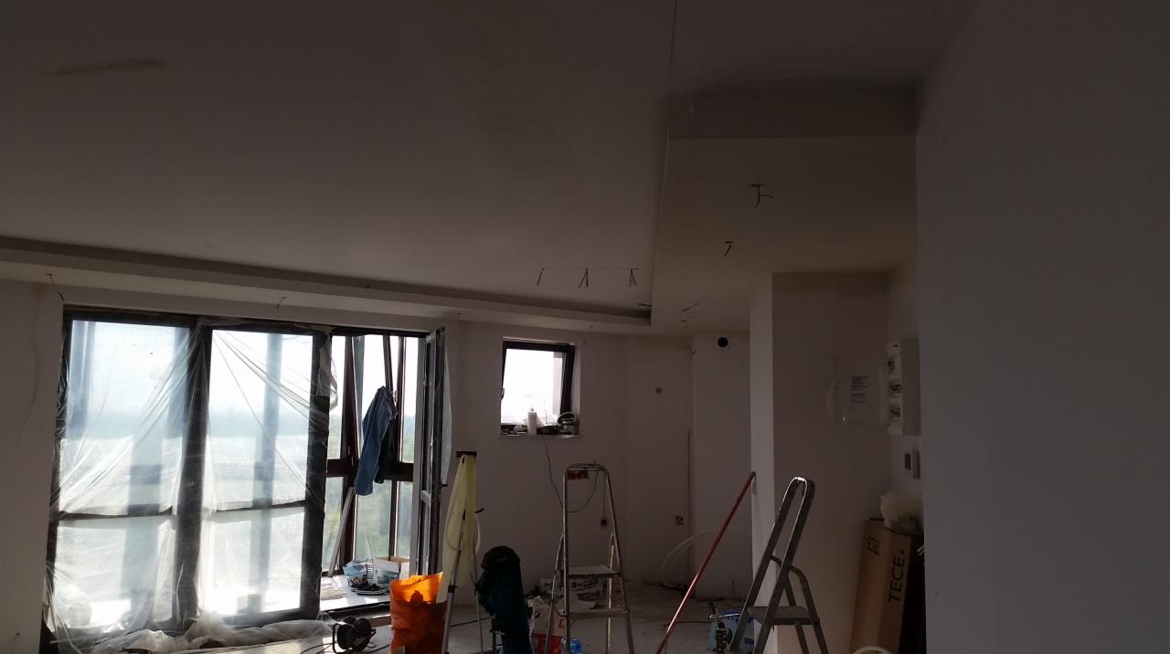 Wnętrze w trakcie prac przygotowawczych: okna zabezpieczone folią, drabiny, zakurzona podłoga, widoczne ślady po instalacjach na suficie.