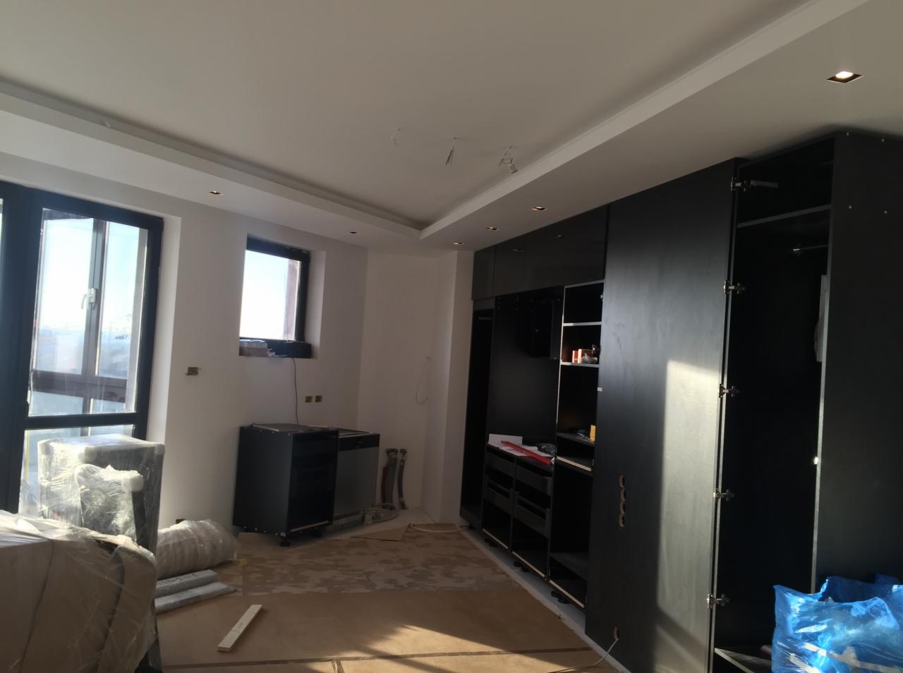 Pokój w trakcie prac wykończeniowych: widoczne okna, czarne szafy w zabudowie, podłoga zabezpieczona folią, elementy wyposażenia w folii ochronnej.